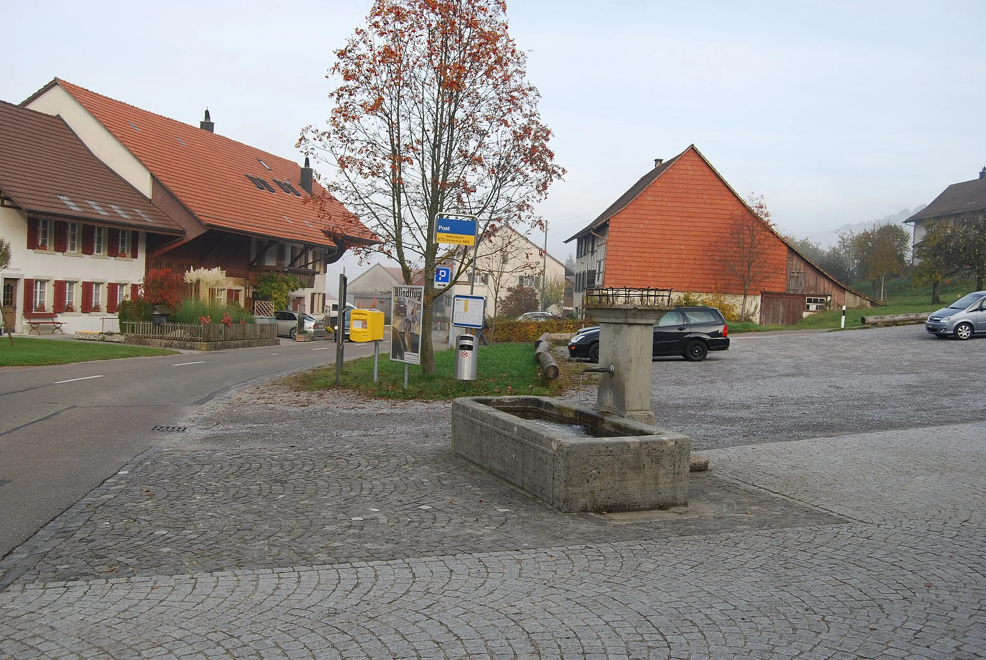 Dorfbrunnen auf einem gepflästerten Platz in Buch am Irchel mit Bauernhäusern im Herbst