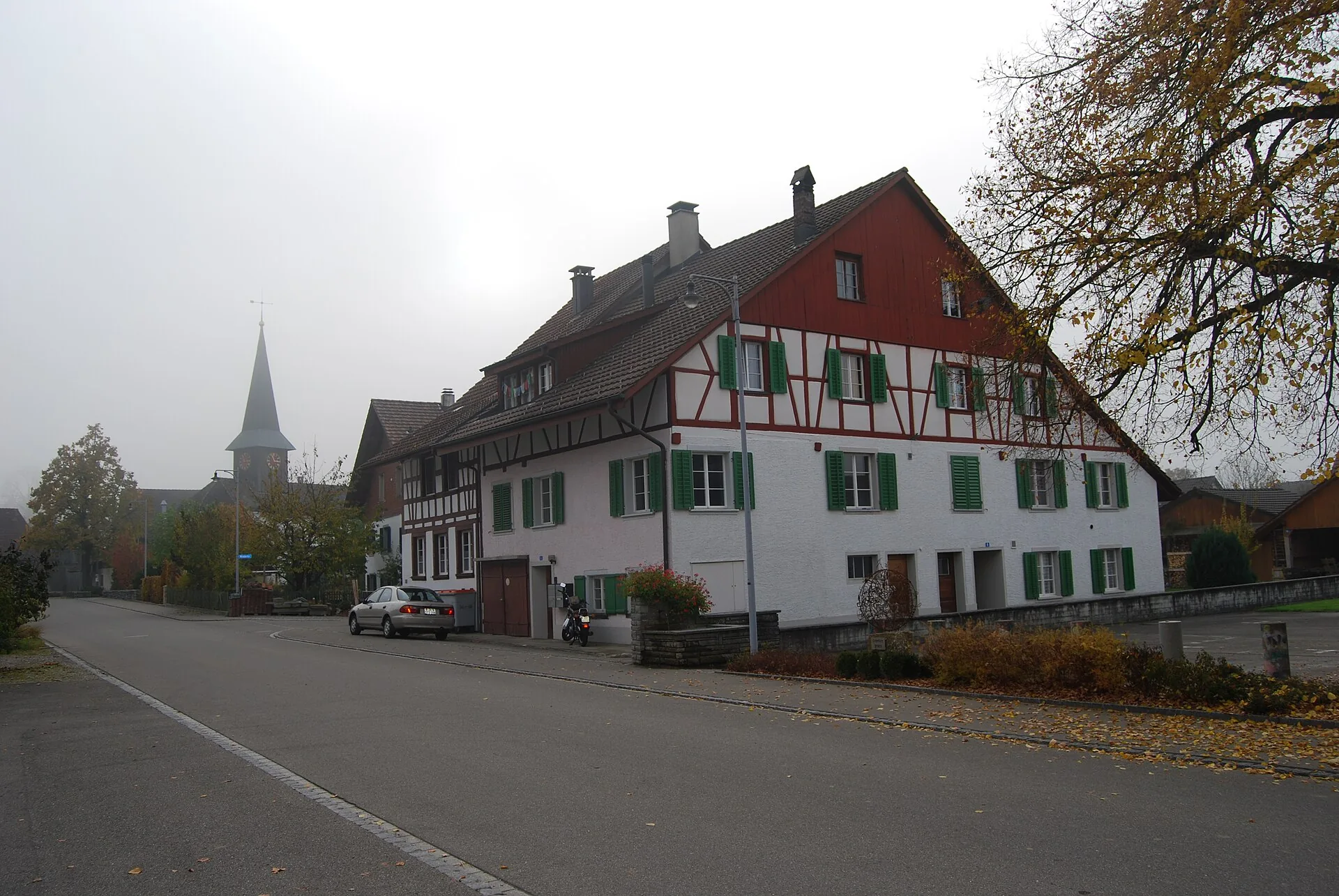 Dorfstrasse in Dorf ZH mit grossem rot-weissem Fachwerkhaus und Kirchturm im Herbstnebel