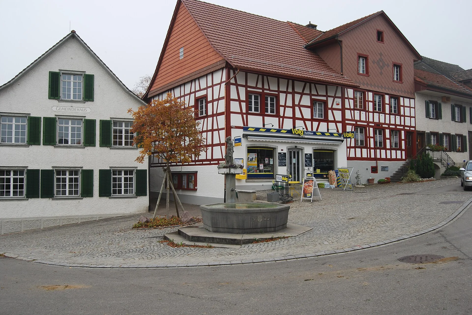 Dorfbrunnen vor einem rot-weissen Riegelhaus im Zentrum von Truttikon, daneben das Gemeindehaus